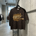 Brązowy, luźny T-shirt z ciemnym wzorem pantery i dużym złotym napisem w stylu graffiti 'SPREAD THE VIBE' na piersi, wiszący na czarnym wieszaku w nowoczesnym sklepie odzieżowym o industrialnym wystroju. Koszulka jest prezentowana na metalowej rurce na tle surowej, betonowej ściany. W tle widać inne wieszaki z ubraniami, betonową podłogę i duże okno.