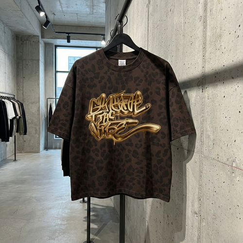 Brązowy, luźny T-shirt z ciemnym wzorem pantery i dużym złotym napisem w stylu graffiti 'SPREAD THE VIBE' na piersi, wiszący na czarnym wieszaku w nowoczesnym sklepie odzieżowym o industrialnym wystroju. Koszulka jest prezentowana na metalowej rurce na tle surowej, betonowej ściany. W tle widać inne wieszaki z ubraniami, betonową podłogę i duże okno.