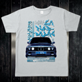 Szara koszulka t-shirt z grafiką BMW E30 w stylu drift glitch na biurku z ciemnego drewna i stalowymi okuciami.