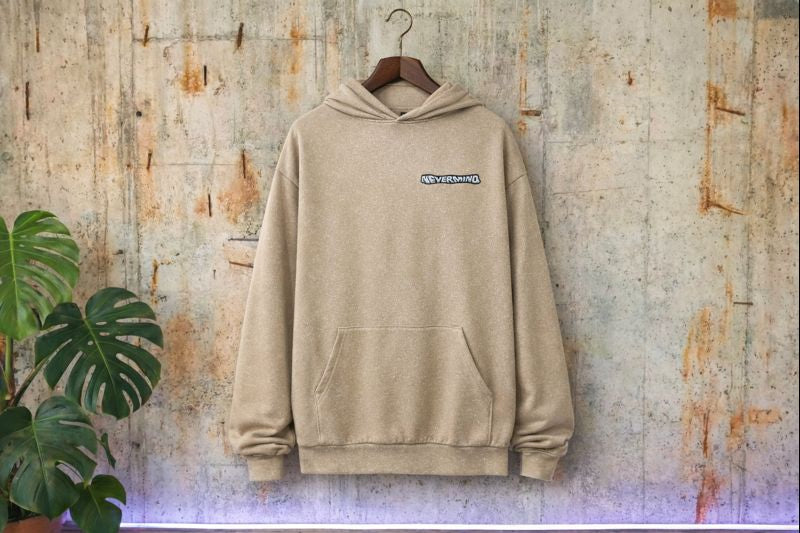 Beżowa bluza z kapturem typu boxy fit z haftowanym logo NEVERMIND, wisząca na drewnianym wieszaku na tle surowego betonu z rośliną.