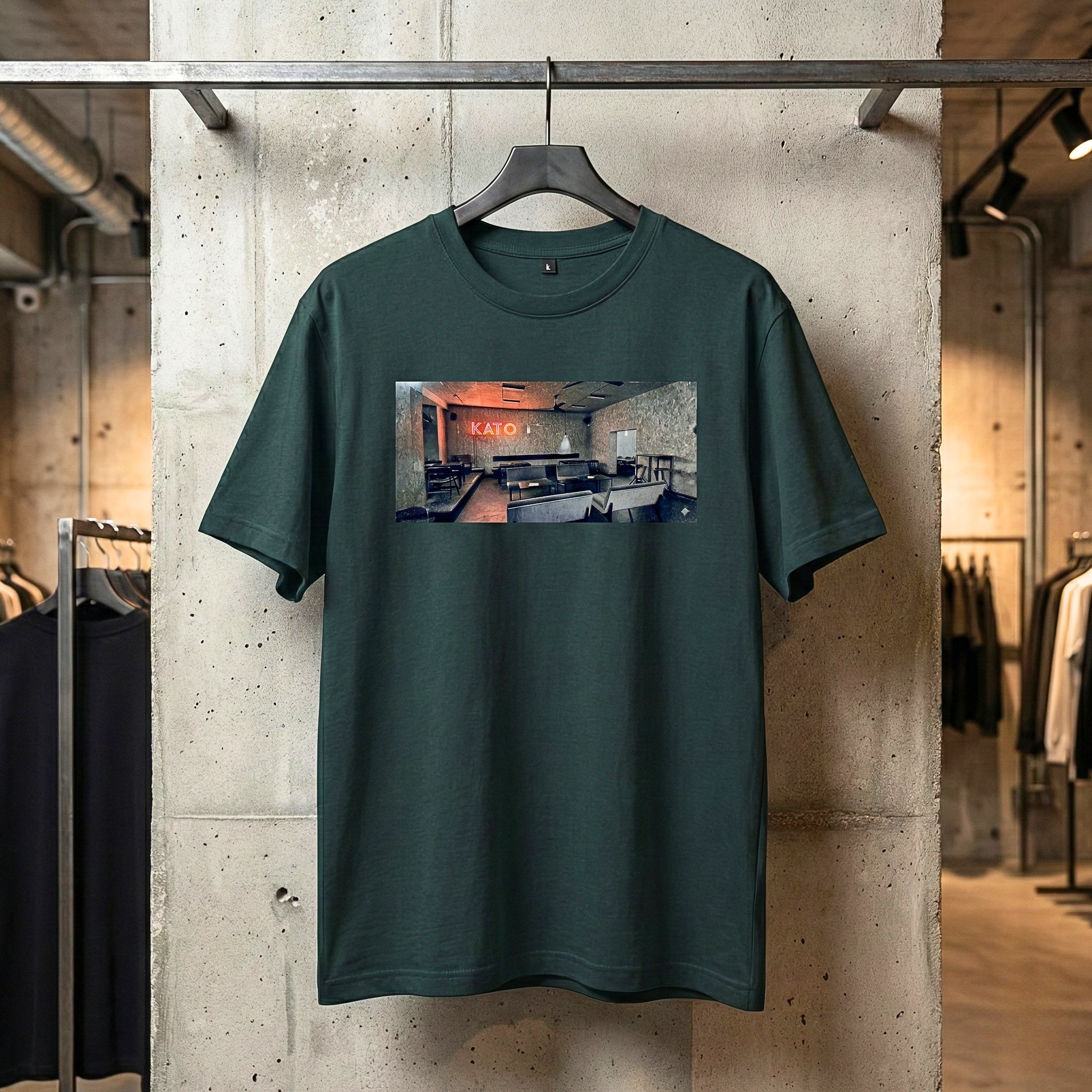 Ciemnozielona koszulka streetwear marki Generate The Vibe wisząca na wieszaku na tle surowej, betonowej ściany. Na froncie t-shirtu znajduje się prostokątny nadruk w mrocznym stylu glitch art, przedstawiający wnętrze lokalu z wyraźnym, czerwonym neonem KATO.